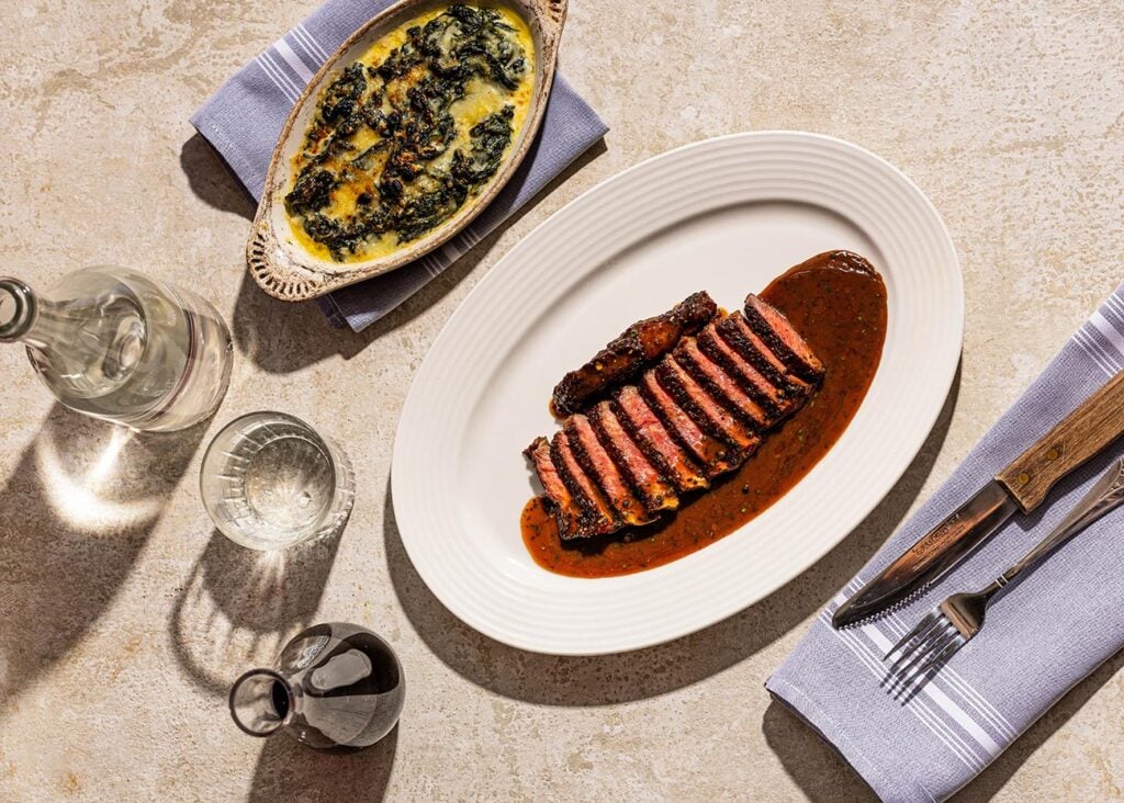 A plate of medium-rare steak and a bowl of baked spinach sit on a restaurant table.