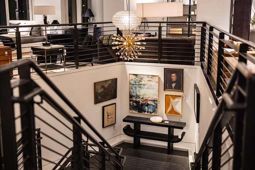 A staircase in the modern furniture store CB2 showing black railings, art on the walls, light fixtures, and contemporary furniture.