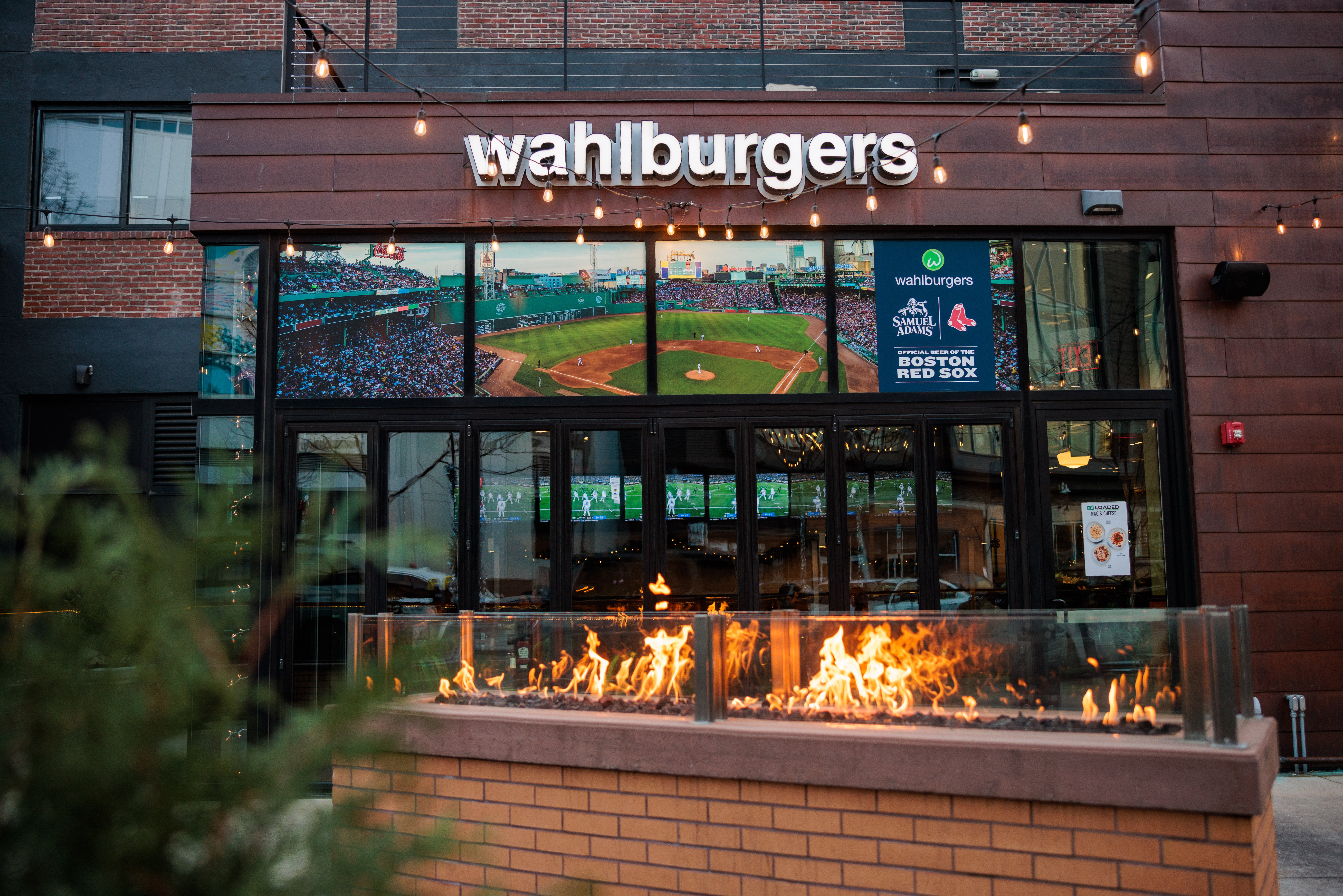 An exterior of the Wahlburgers restaurant showing an inviting outdoor fire pit; inside of the space shows many TV screens displaying the Red Sox game.