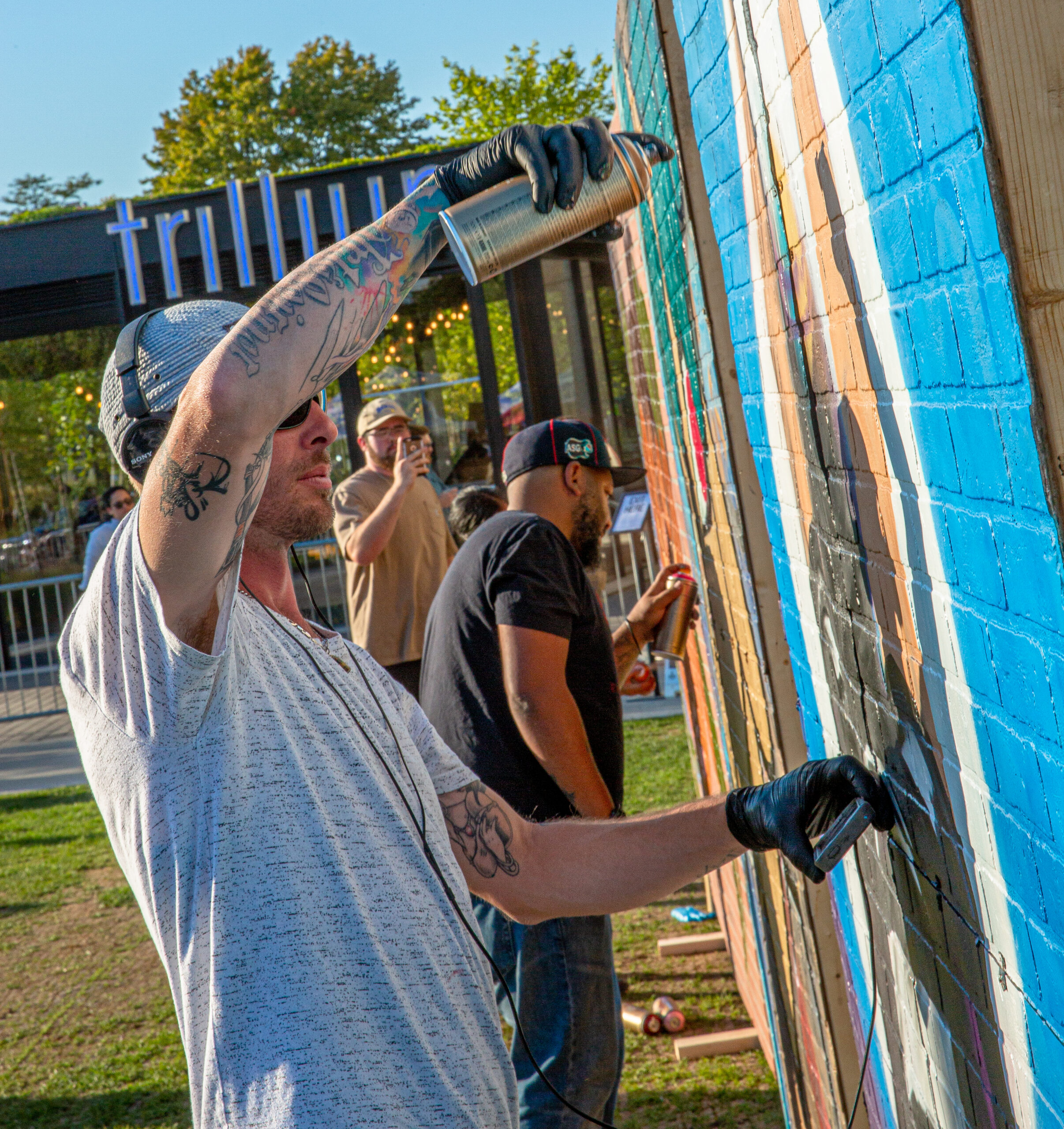 Two graffiti artists paint a wall outside of Trillium brewery while another man snaps a photo of them.